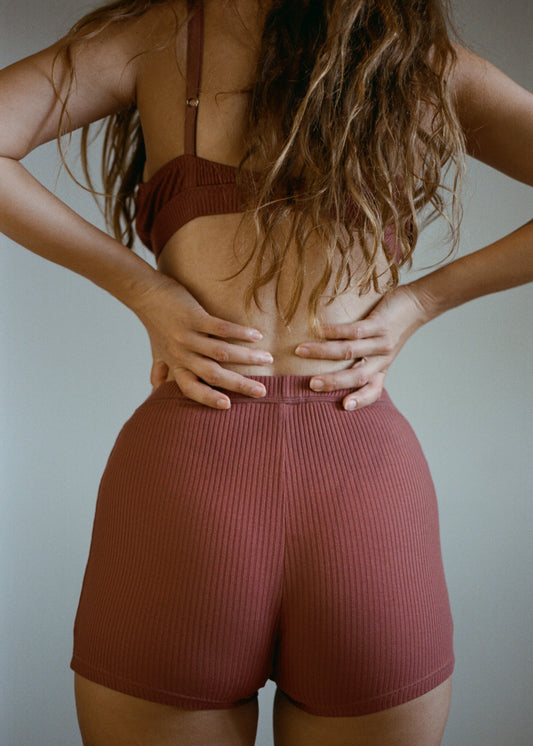 A portrait image of a women with long wavy brown hair with her back to the camera. She is wearing the  Maggie Short in Clay by Juem with a matching bra, and her hands are on her hips. She is standing in front of a white wall.