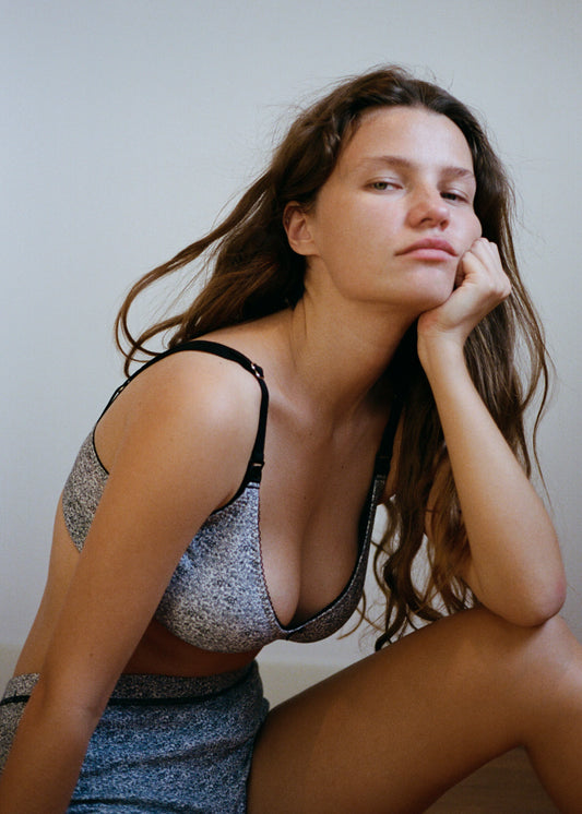 A woman is sitting on the ground leaning forward with her arm resting on her bent knee, and her hand in her chin. She is looking directly into the camera, and has a neautral face, and her long brown hair is falling over her shoulders. She is wearing the Maggie Short in Midnight Floral by Juem with a bra, in front of a white wall.