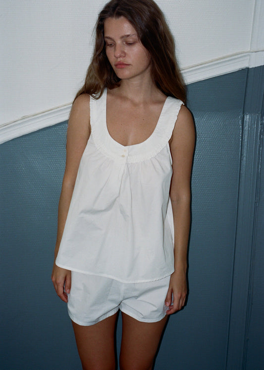 A woman with brown, wavy hair is gazing away from the camera with her arms by her side. She is wearing the white Joey cotton top and Joey cotton shorts set by Juem. She is standing in front of a white and blue wall.