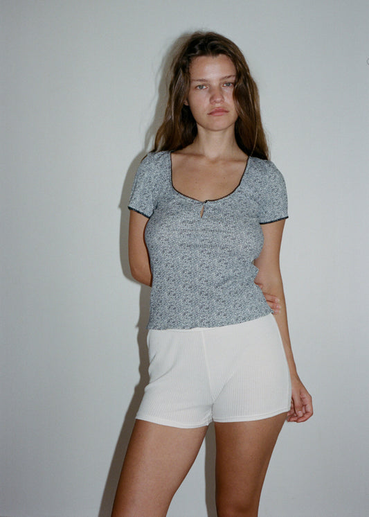 A woman is standing in front of a light coloured wall, with brown hair. One arm is crossed behind her back. She is looking into the camera, wearing the Effie Tee in Midnight Floral by Juem paired with a white microshort.