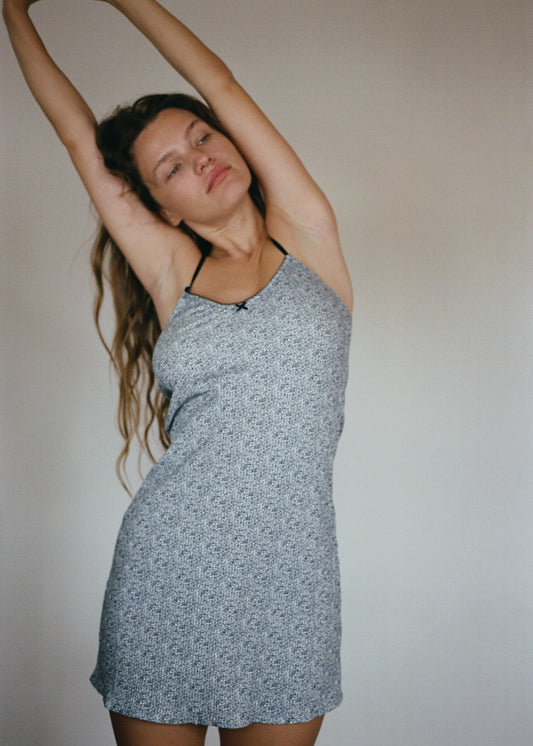 A woman with long, wavy brown hair is standing in front of a white wall with her hands stretched above her head. She is looking away from the camera. She is wearing the Lola Mini Dress in Midnight Floral by Juem.