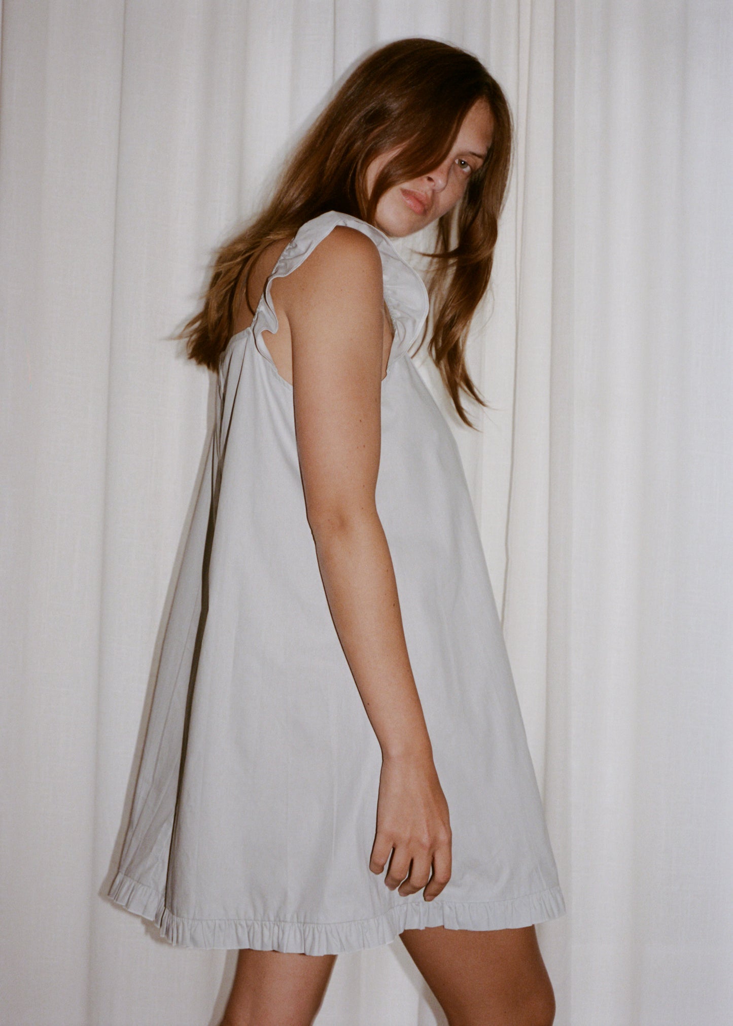 A woman with long brown hair stands sideways in front of white curtains, wearing the 0 Jonni Dress in powder blue—a loose, sleeveless organic cotton dress with ruffled edges—looking at the camera with a neutral expression.