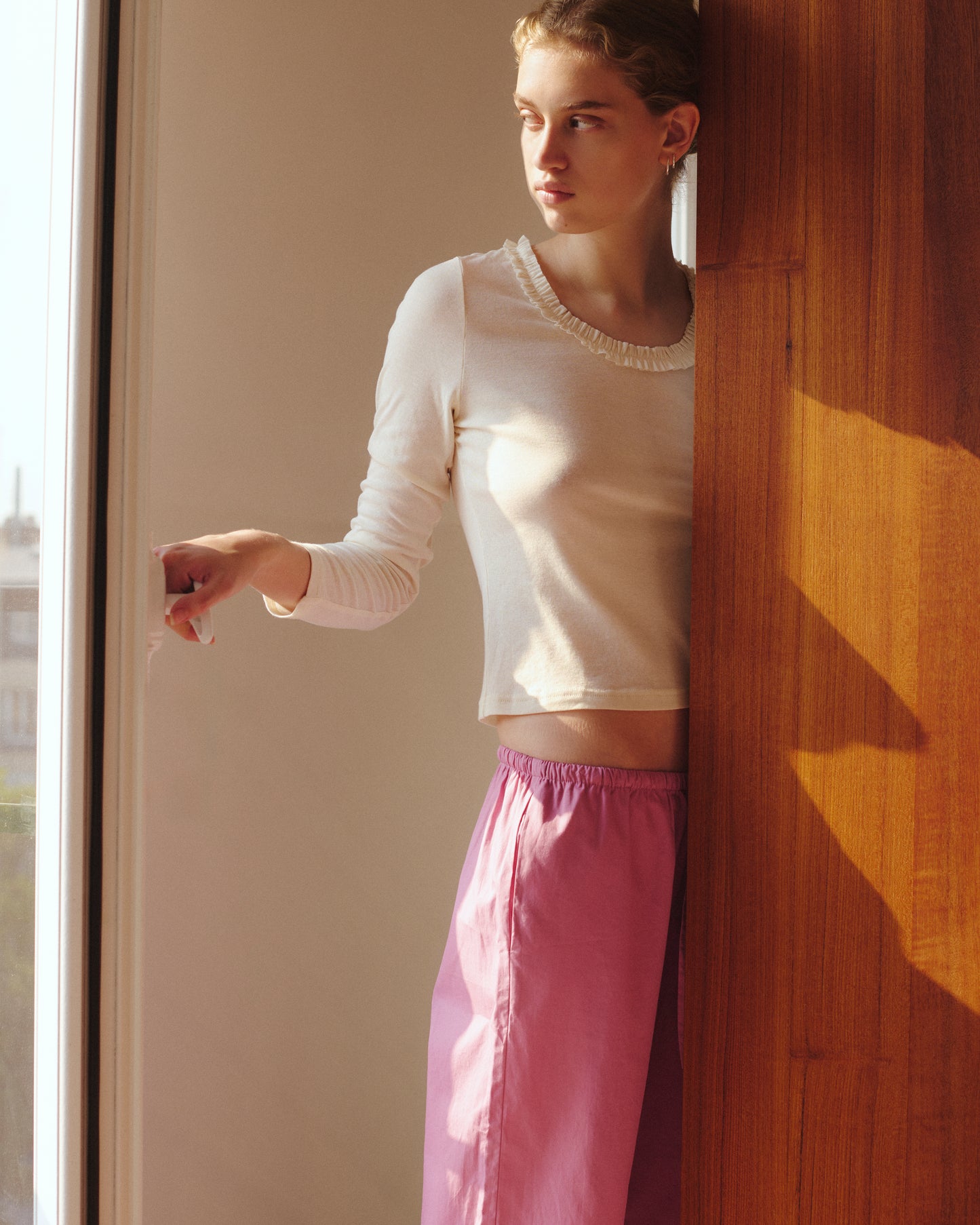 Wearing the Ellery Capri in Mauve by 0 and a white long-sleeve top, a person stands by a wooden wall, gazing out of a window as soft sunlight fills the scene.