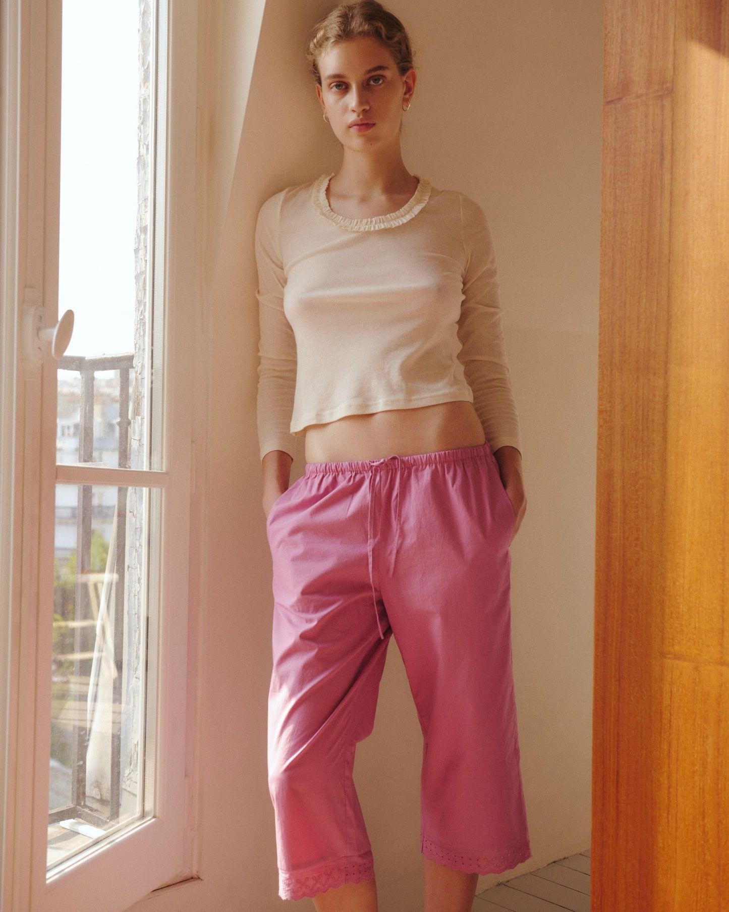 A person stands by a window in soft natural light, gazing at the camera while wearing the Claude Long Sleeve - Butter from 0, an organic cotton cream top with a subtle lettuce frill trim, paired with loose pink shorts and hands in pockets.