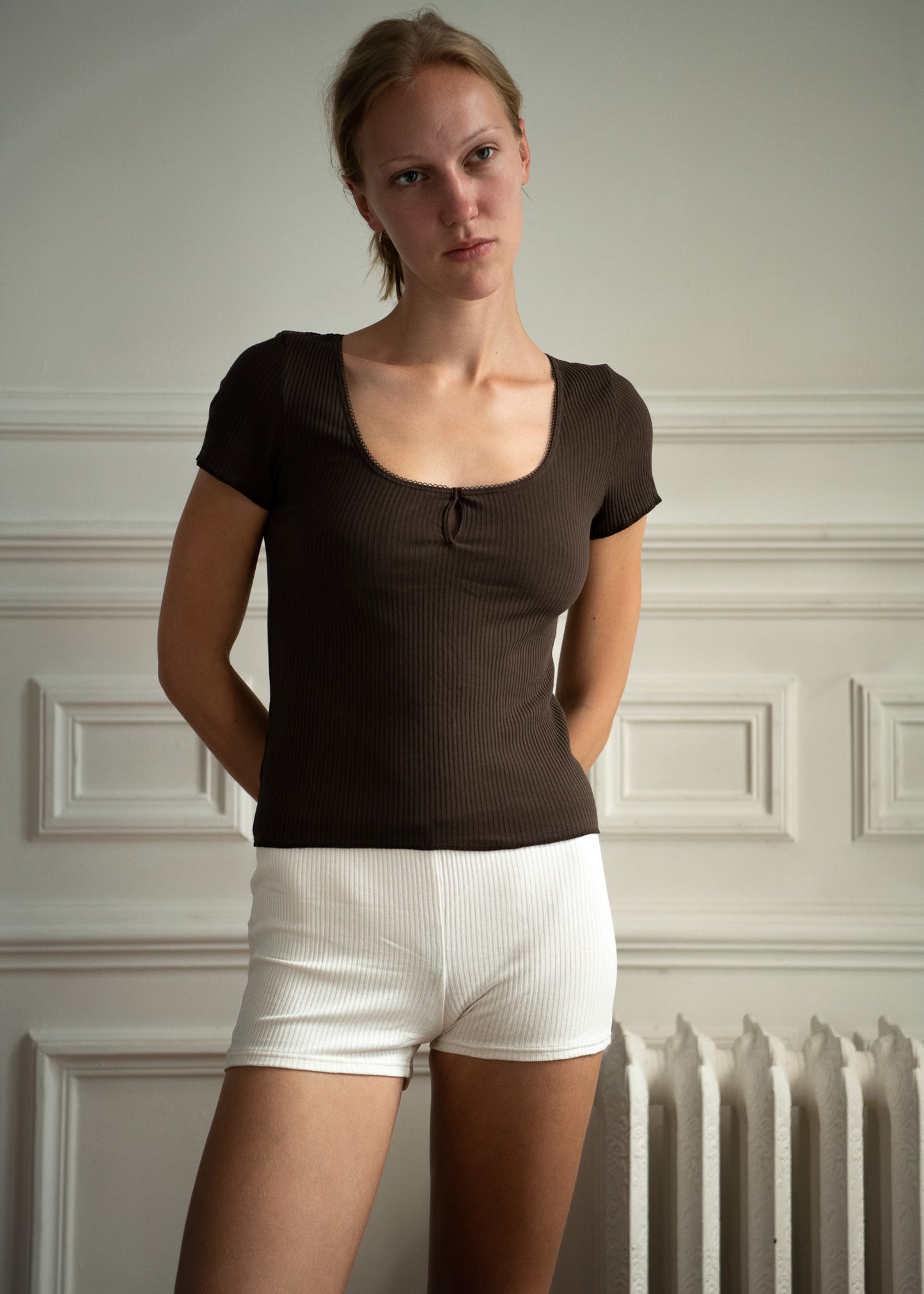 A woman with blonde hair stands against a light wall, wearing Juem Maggie Short in pumice with a chocolate ribbed tee. She has both arms rested on her hips and is looking just off from the camera with a soft and relaxed expression.