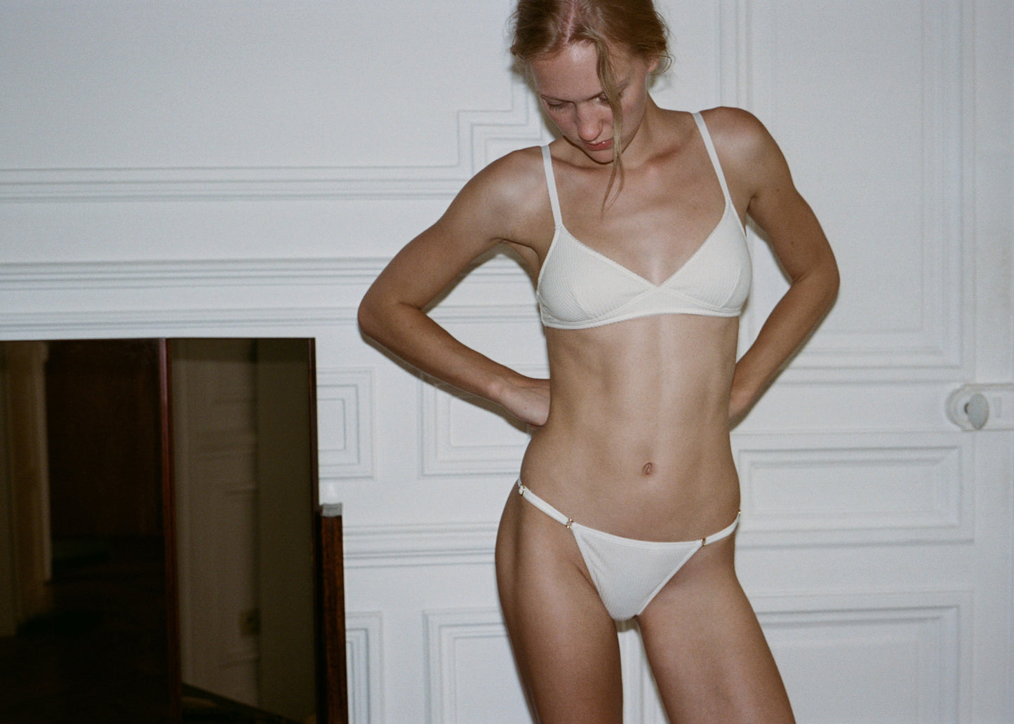 A woman with blonde hair stands against a light wall wearing Juem Harri G String in pumice with a matching white bralet top. She has both her hands resting on her hips and is looking downwards towards the ground