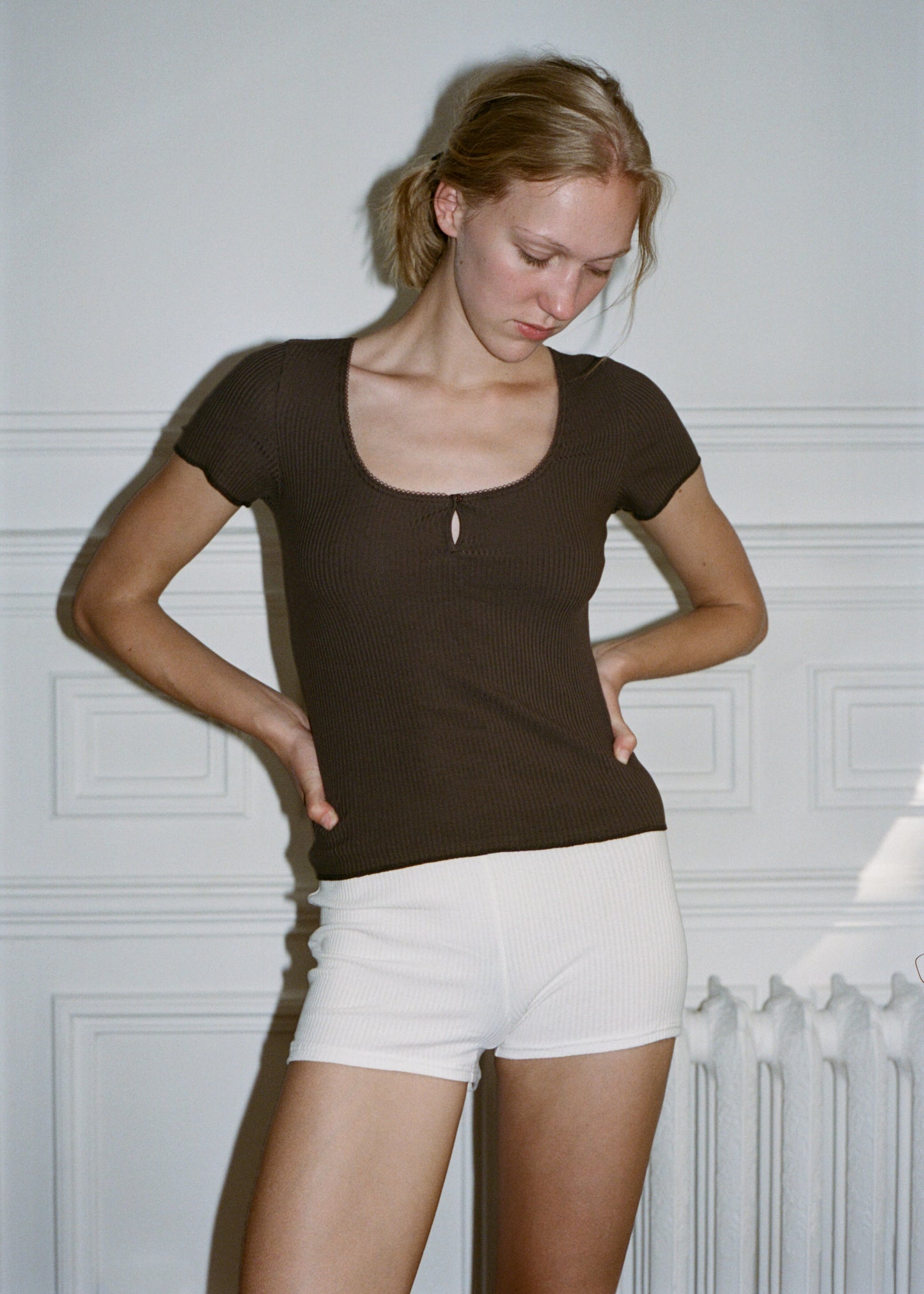 A woman with blonde hair stands against a light wall, wearing Juem Maggie Short in pumice with a chocolate ribbed tee. She has both arms rested on her hips and is looking down and away from the camera with a soft and relaxed expression.