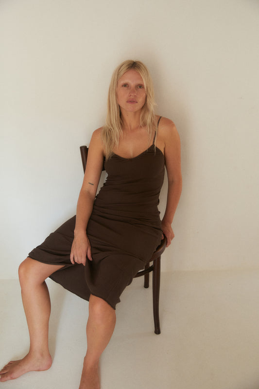 Woman with long blonde hair sitting barefoot on a wooden chair against a plain off-white wall, wearing a fitted, ribbed brown midi dress with thin straps. Her expression is calm and natural in the softly lit, minimal setting.
