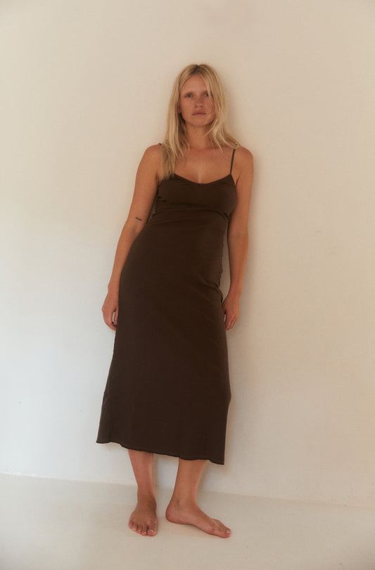 Woman with long blonde hair barefoot standing against a plain off-white wall, wearing a fitted, ribbed brown midi dress with thin straps. Her expression is calm and natural in the softly lit, minimal setting.