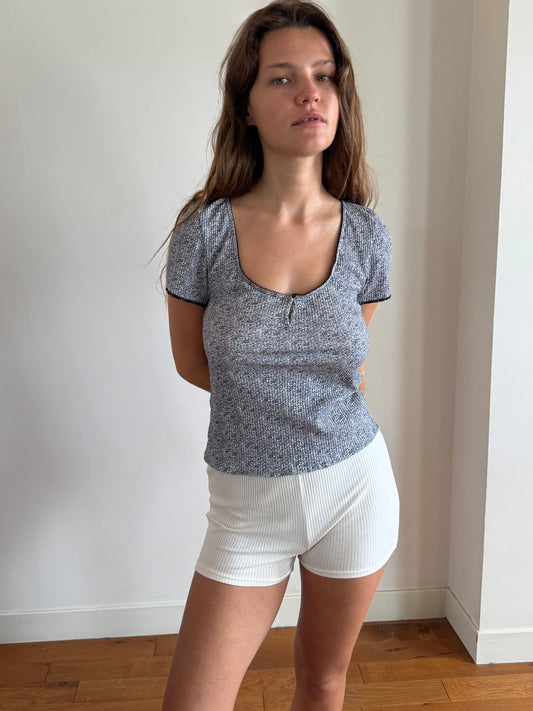 A girl is standing in front of a white wall looking into the camera with her arms crossed behind her back. She wears the Effie Tee in Midnight Floral by Juem with white shorts. She is standing on timber floorboards, and has wavy brown hair.