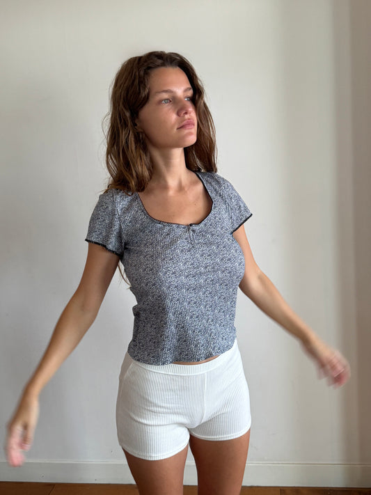 A brunette woman has her arms slightly raised, standing on floorboards in front of a white wall. She is wearing the Effie Tee in Midnight Floral by Juem with white boyleg shorts, and is looking out a window.