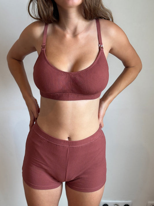 A close up image shows a woman wearing the Maggie Short in Clay by Juem with a bra. Her hands are on her hips, and she is standing in front of a light coloured wall that has a power point. She has brown hair.