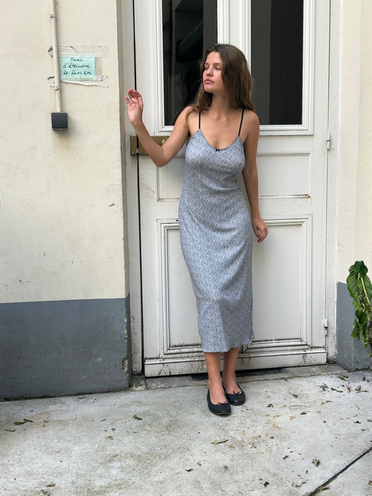 A woman with brown hair is standing in front of a white door, with light coloured walls and a blue skirting. There is a green plant next to her. She is looking to her right with her hand raised in a wave, wearing the Rafi Midi Dress in Midnight Floral by Juem paired with black ballet flats with a bow. She is standing on concrete flooring.