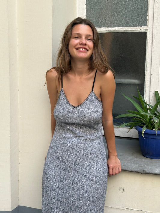 A woman is leaning against a window frame next to a pot plant. She is smiling into the camera and has long brown hair. She is wearing the Rafi Midi Dress in Midnight Floral by Juem. The walls are white in the background.