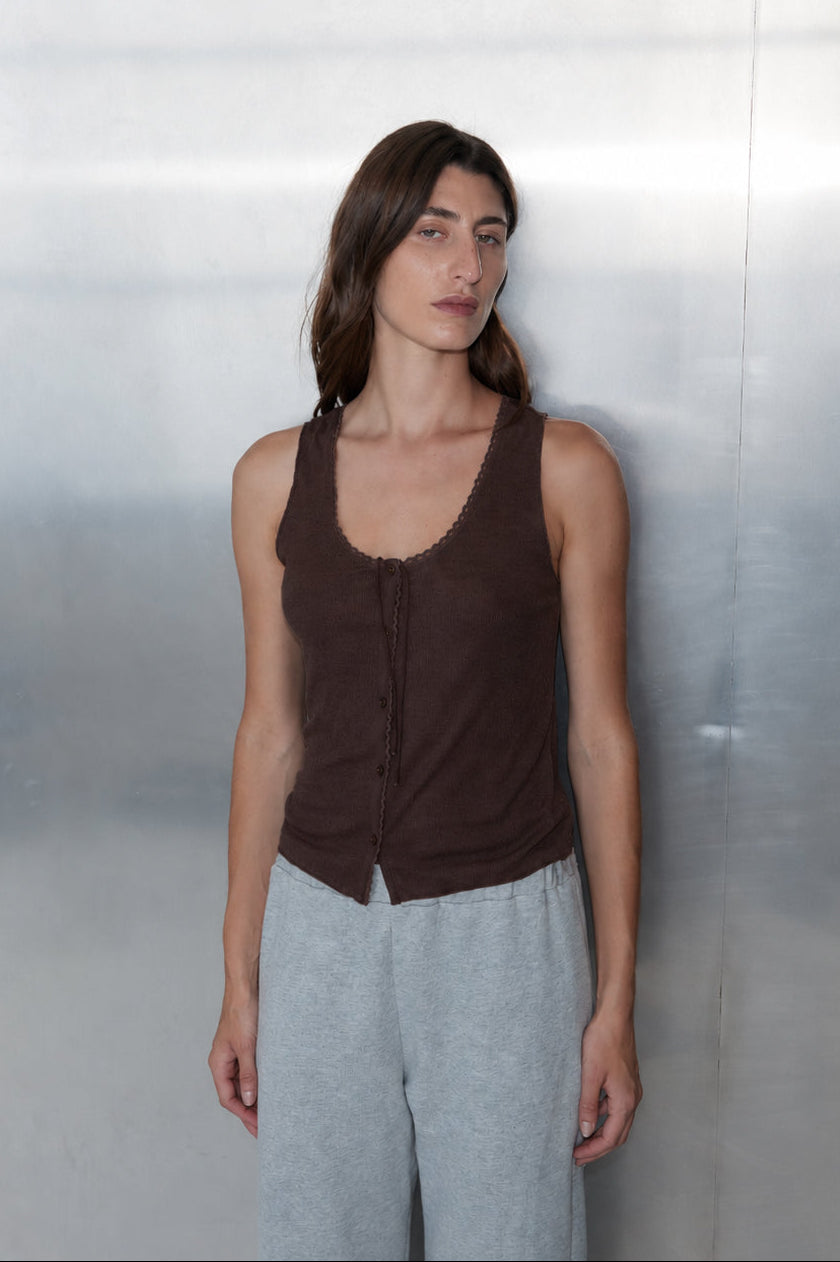 A woman with long brown hair stands against a metallic wall, wearing the 0 Monte Tank in Chocolate with a scoop neckline and light gray pants. She has a neutral expression, arms relaxed at her sides.
