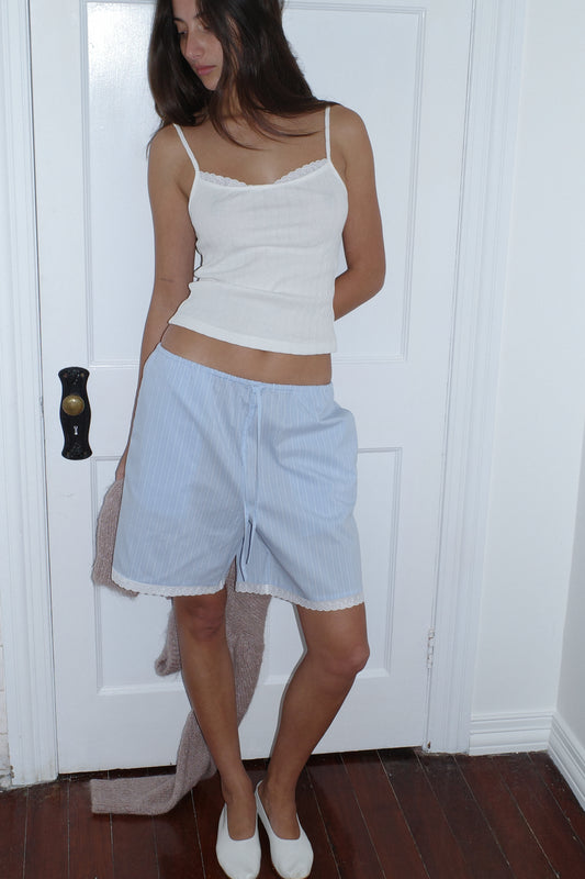 A woman stands indoors on a wooden floor, wearing the 0 Joan Tank in Off White with adjustable straps, light blue drawstring shorts, and white shoes. She holds a beige sweater and stands before a white door.
