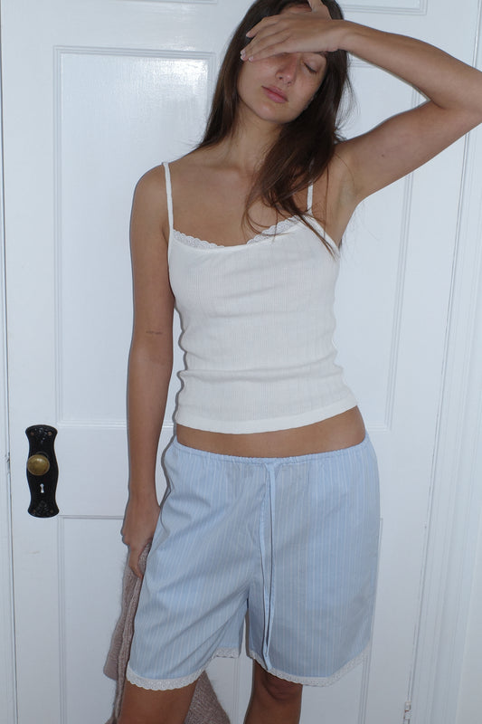 A woman in the Mae Short - Cloud Stripe by 0, featuring light blue stripes and an elastic waistband, stands before a white door with one hand over her eyes, looking tired or frustrated.