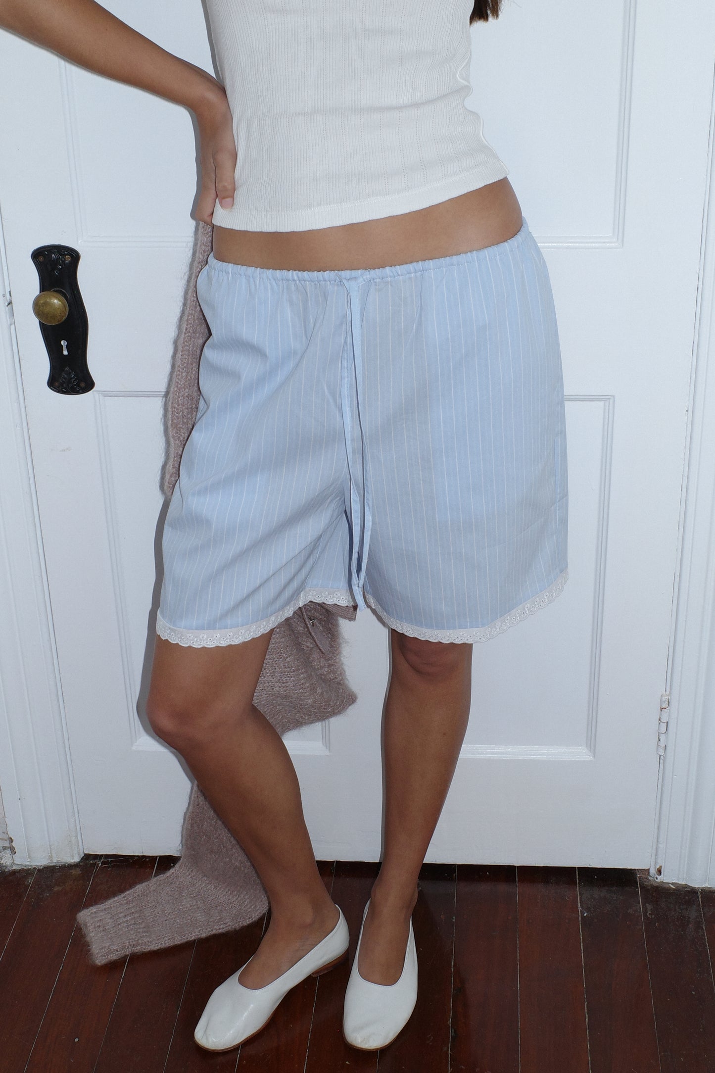 A person stands by a white door on a wooden floor, wearing the 0 Mae Short - Cloud Stripe paired with a white tank top and holding a brown sweater. Their head is not visible in the image.