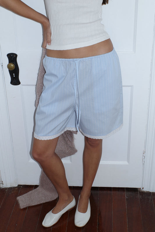 A person stands by a white door on a wooden floor, wearing the 0 Mae Short - Cloud Stripe paired with a white tank top and holding a brown sweater. Their head is not visible in the image.