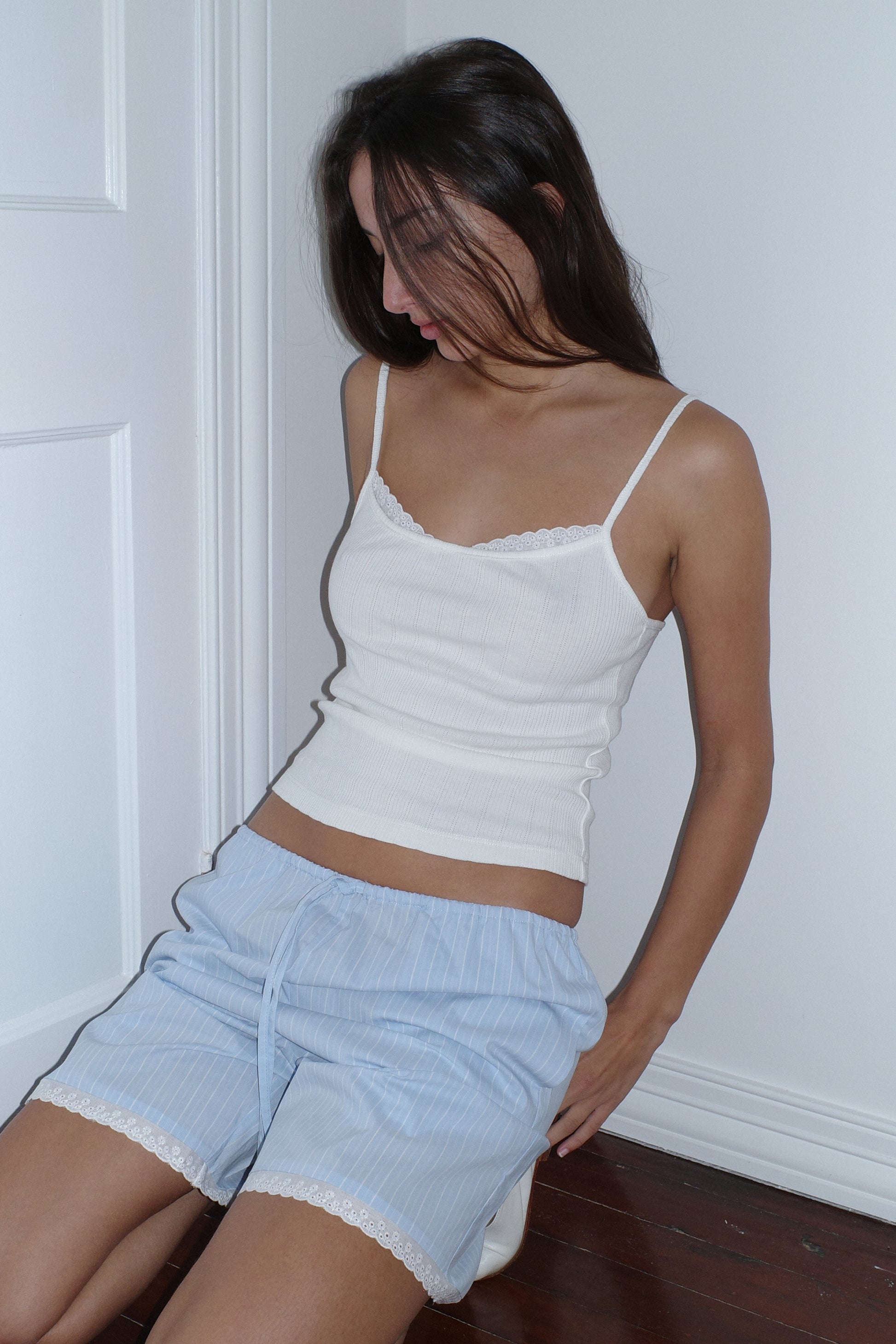A woman with long brown hair sits on a wooden floor against a white wall, wearing a white camisole and the 0 Mae Short - Cloud Stripe, light blue organic cotton shorts with lace trim. Her face is partly hidden by her hair as she looks down.