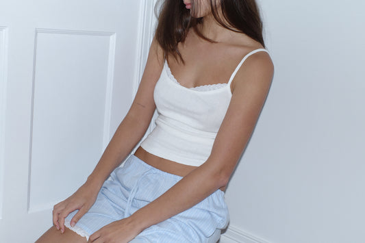 A woman wears the 0 Joan Tank in Off White, paired with light blue striped shorts, sitting against a white wall with her face partially out of frame and brown hair over her shoulder.