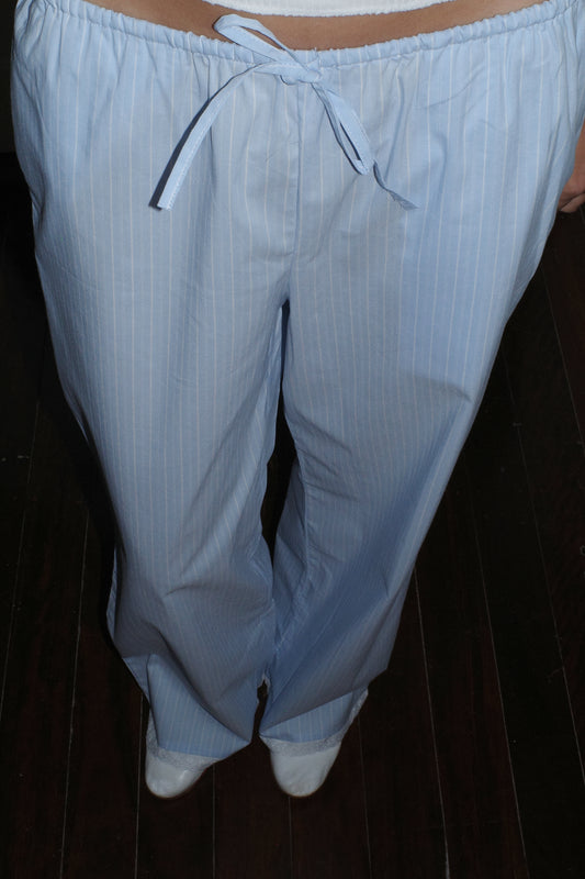 A person wearing the Josephine Pant - Cloud Stripe by 0, featuring light blue pinstripes, organic cotton fabric, drawstring waist, and relaxed fit, stands on a dark wooden floor with only the pants and part of the shoes visible.