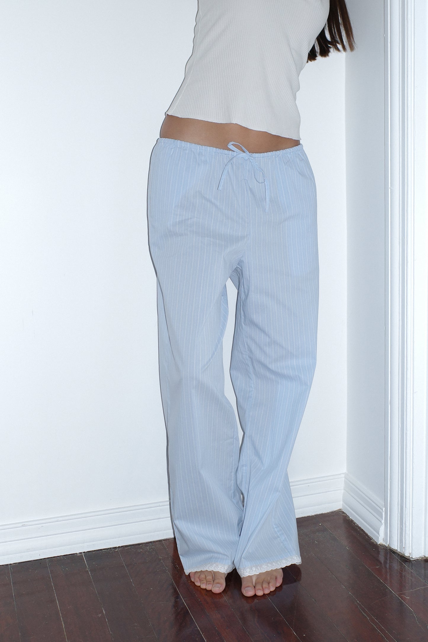 A person stands barefoot on a dark wood floor, wearing the Josephine Pant - Cloud Stripe by 0 and a white sleeveless top, leaning against a white wall with their head out of frame.