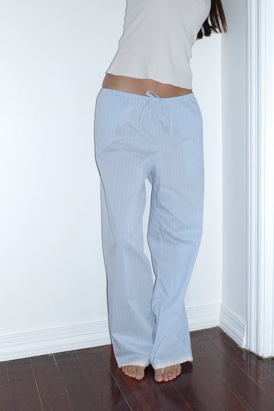 A person stands barefoot on a dark wood floor, wearing the Josephine Pant - Cloud Stripe by 0 and a white sleeveless top, leaning against a white wall with their head out of frame.