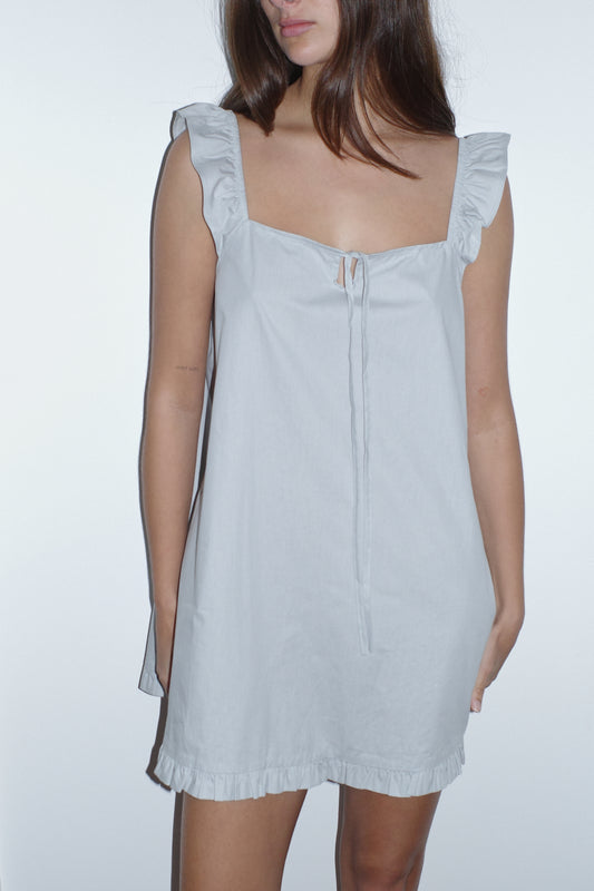 A woman stands against a white background wearing the 0 Jonni Dress in powder blue, an organic cotton mini dress with ruffled shoulders and hem, plus a tie at the neckline. Her face is partially out of frame.