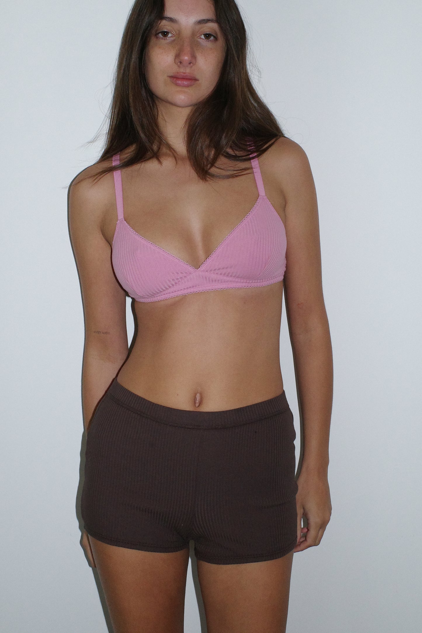 A woman with long brown hair and a neutral expression stands against a white wall, wearing the 0 Maggie Short in Chocolate and a pink bralette.