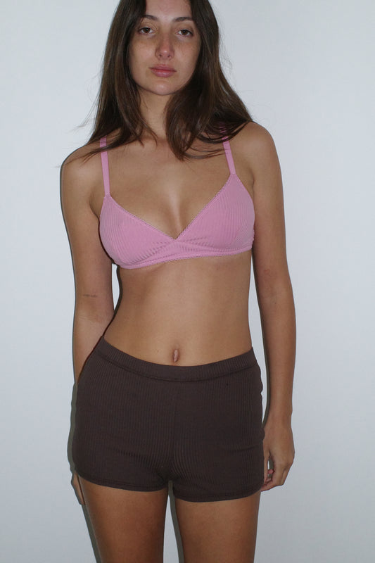 A woman with long brown hair and a neutral expression stands against a white wall, wearing the 0 Maggie Short in Chocolate and a pink bralette.