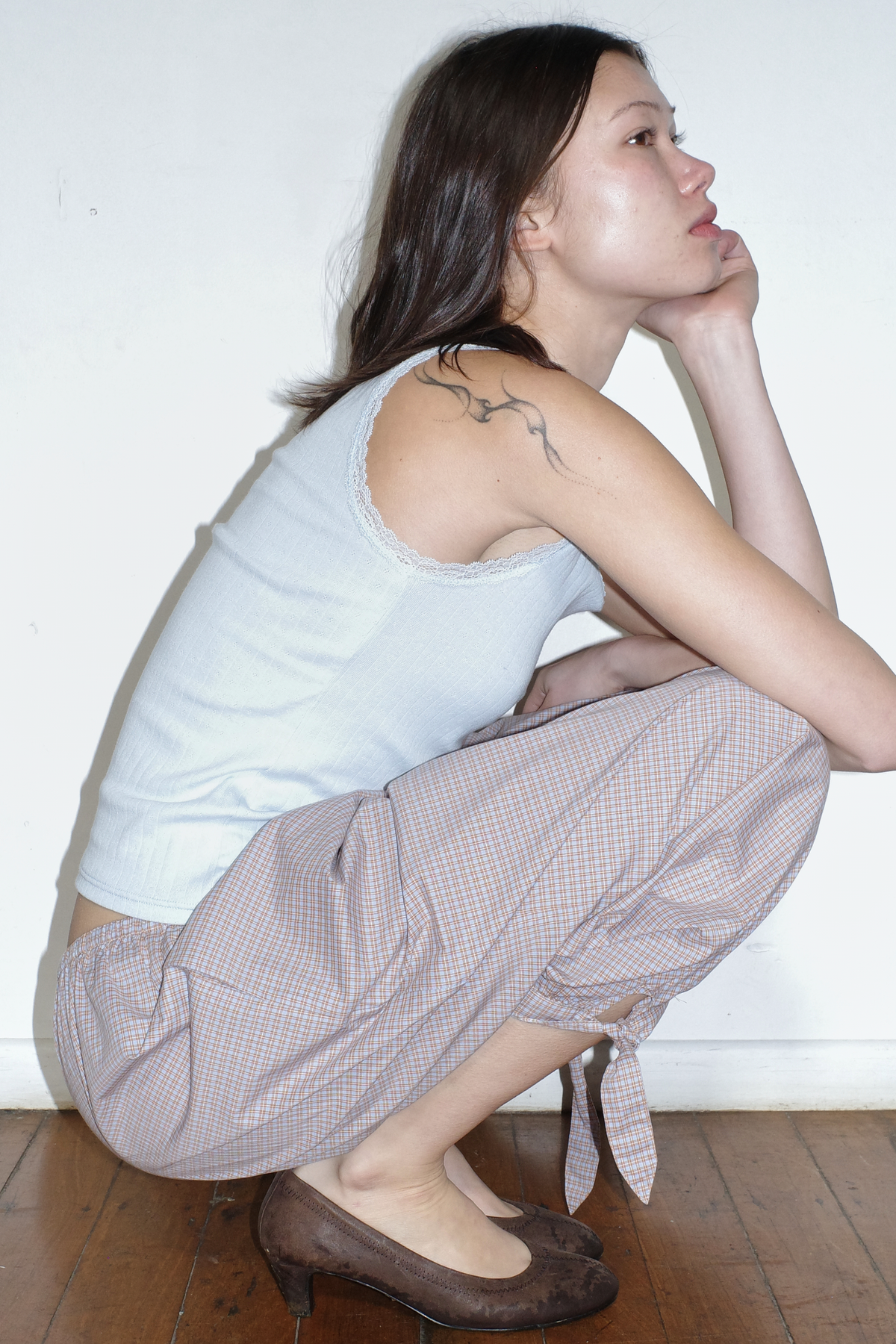A model crouches in profile against a white wall, hand to chin, wearing the Betty Singlet - Glacier Blue by Juem, a lace-trimmed pointelle knit tank, styled with the Freya Pant in Blue Tan Check and brown heels.