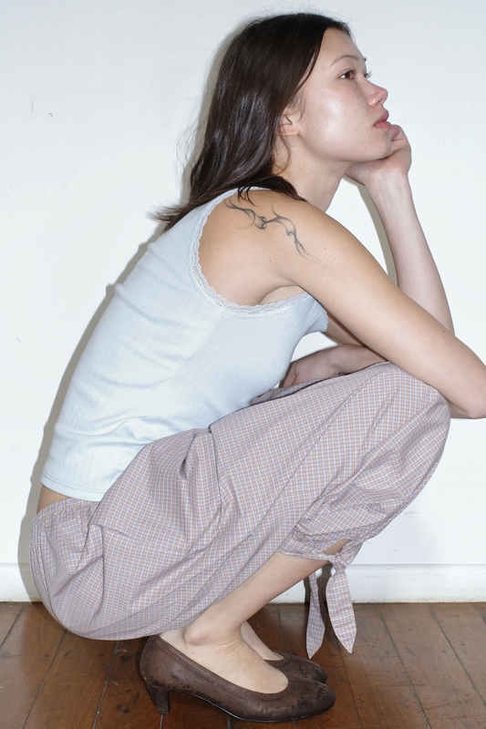 A model crouches in profile against a white wall, hand to chin, wearing the Betty Singlet - Glacier Blue by Juem, a lace-trimmed pointelle knit tank, styled with the Freya Pant in Blue Tan Check and brown heels.