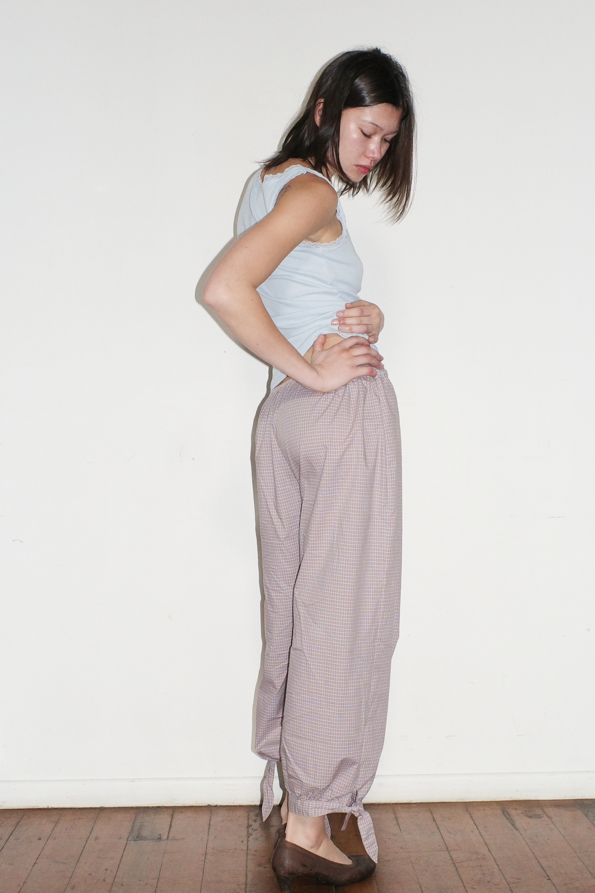 A model stands in profile against a white wall, looking down, wearing the Betty Singlet - Glacier Blue by Juem, a lace-trimmed pointelle knit tank, styled with the Freya Pant in Blue Tan Check.