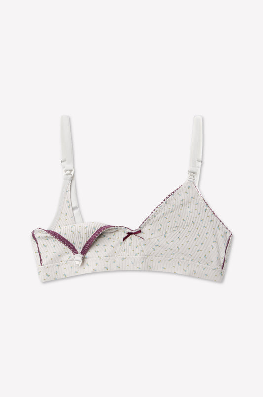 Introducing the Paloma Maternity Bra - Blue Floral by 0: a soft organic cotton maternity bra featuring thin straps, purple trim, and a delicate front bow. Its small green and yellow floral print adds a charming touch. Designed with easy-to-use nursing clips—the left cup is shown unclipped and folded down for convenient breastfeeding access.