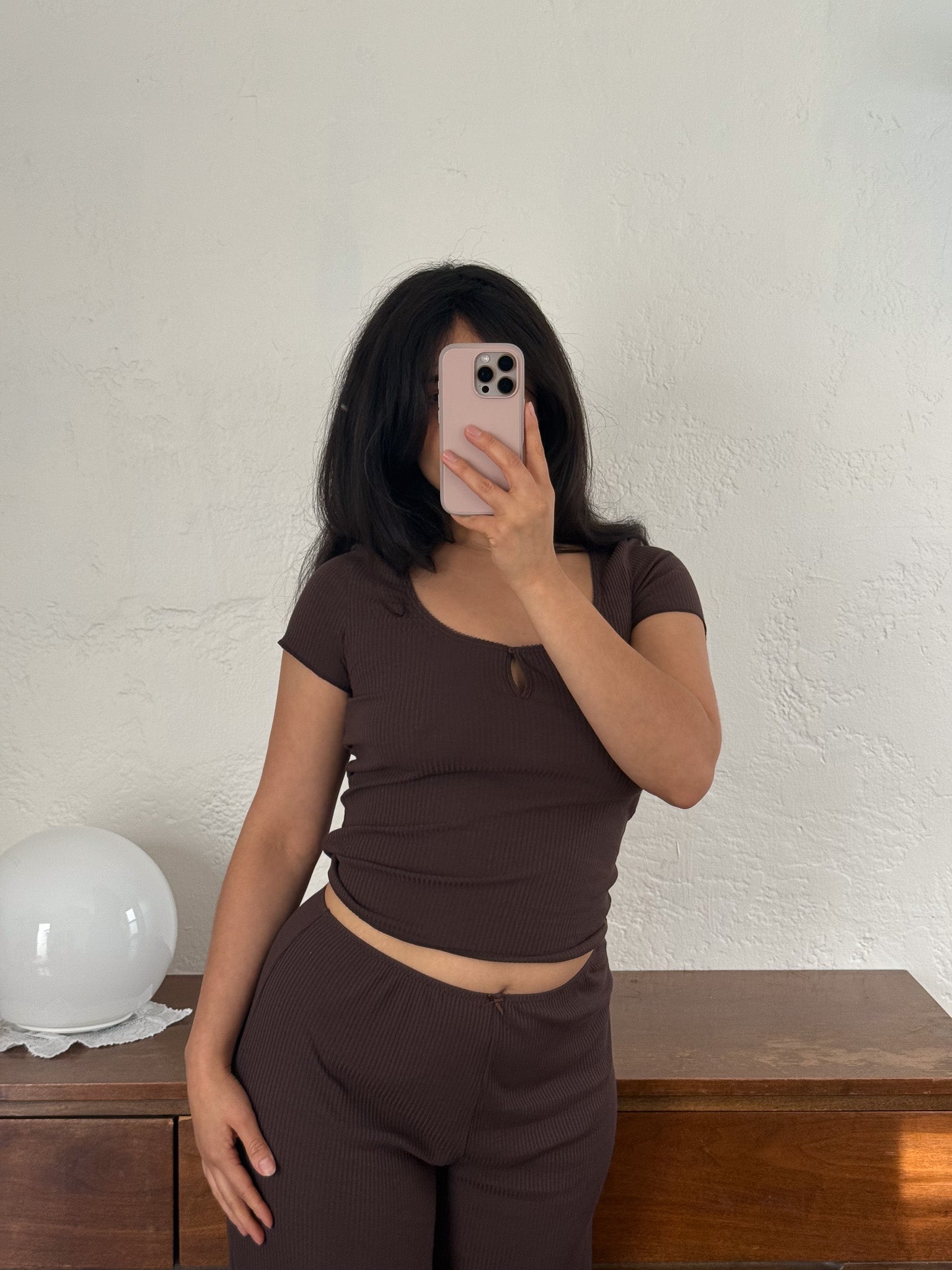 A person with long dark hair wears the 0 Effie Tee in Chocolate, paired with matching pants. They stand indoors, holding a phone up to their face in front of a textured white wall and a wooden dresser topped with a white lamp.