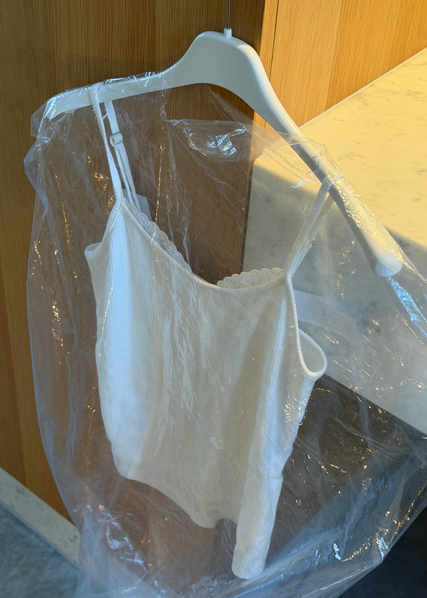The Joan Tank - Off White by 0, a sleeveless ribbed camisole with adjustable straps, hangs on a white hanger under a clear garment bag near a wooden wall and white marble countertop.