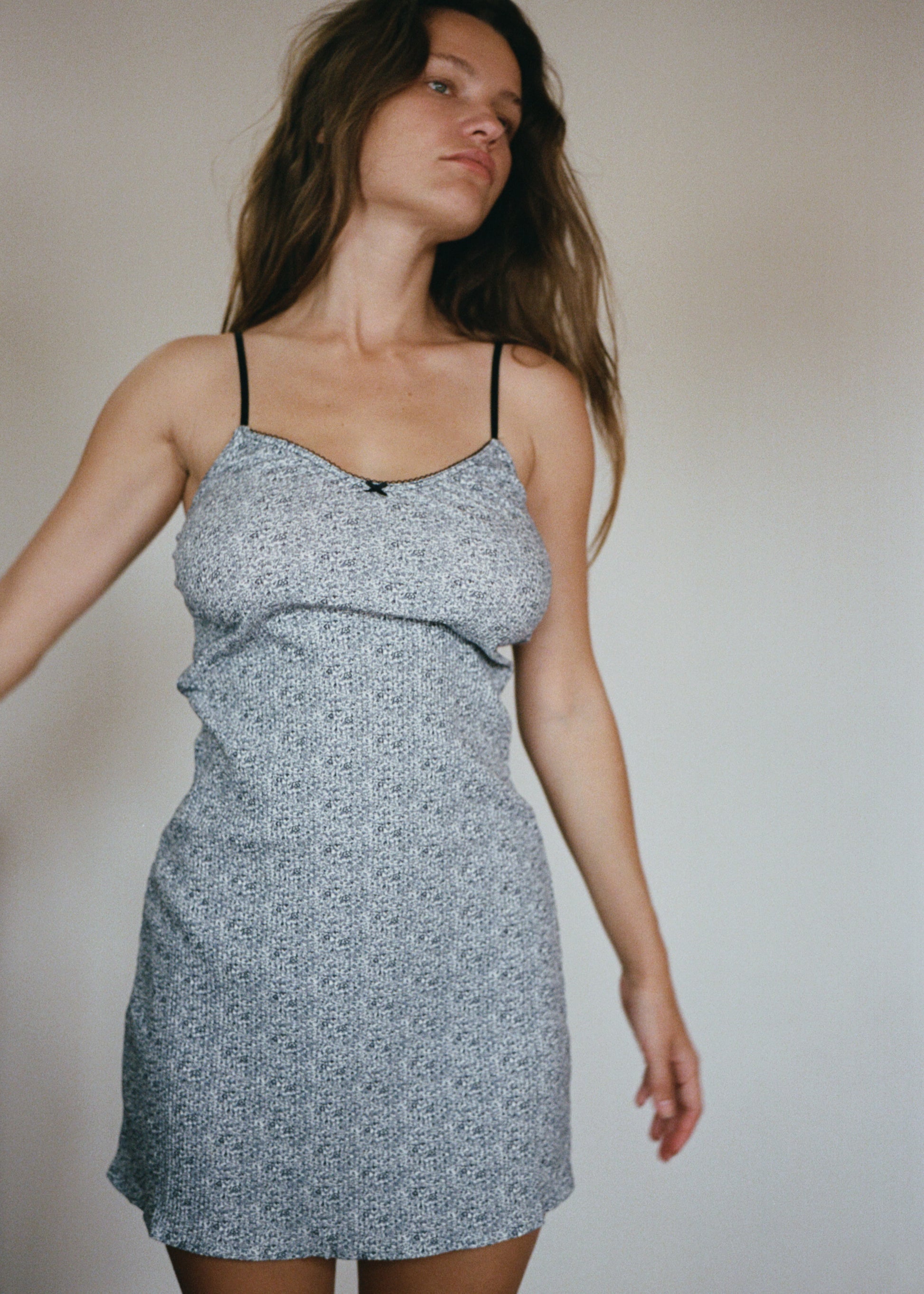 A woman is wearing the Lola Mini Dress in Midnight Floral by Juem, she is brunette and has her hands placed by her side,  looking to the left. There is a light coloured wall behind her. 