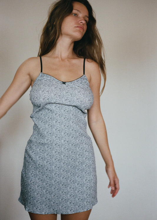 A woman is wearing the Lola Mini Dress in Midnight Floral by Juem, she is brunette and has her hands placed by her side,  looking to the left. There is a light coloured wall behind her. 
