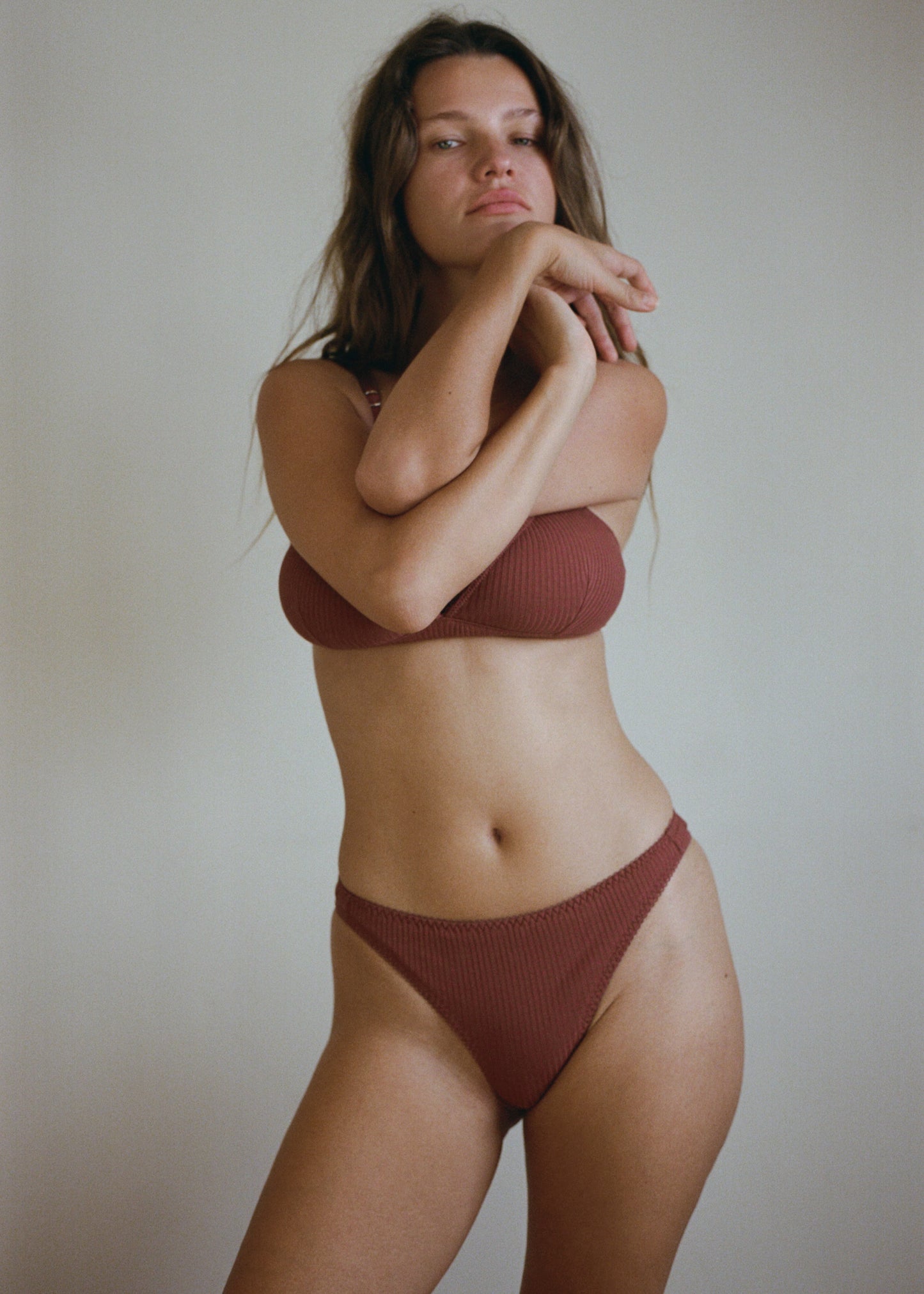 A woman with brown hair is standing in front of a white wall. She is wearing the Lera Brief in Clay by Juem with a bra. Her arms are crossed in front of her with her hands held up to her face. 