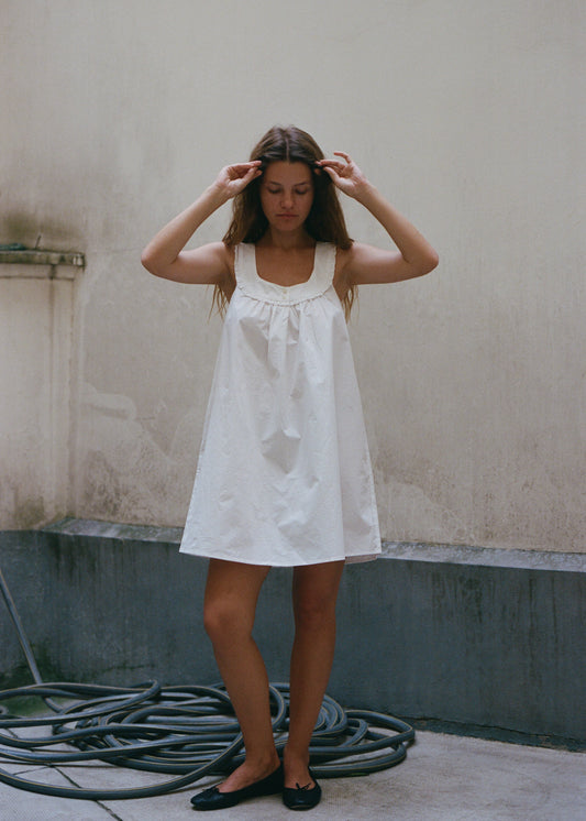 A woman with long brown hair wears the white cotton Olivia dress by Juem. She is standing against a plain white wall with a green skirting, and a hose on the ground. She wears black ballet flats with a bow, and is pushing her hair back with her hands whilst looking down towards the ground. 