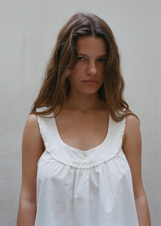 A portrait image of a woman who is staring directly into the camera, she has wavy long hair and is wearing the white cotton Olivia Dress by Juem. She is standing against a plain white wall. 