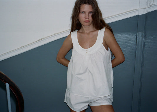 A woman with brown hair is wearing the  white sleeveless top and matching shorts Joey set by Juem. She is standing against a blue and white wall with her arms behind her back, and gazing into the camera with a neutral expression. 