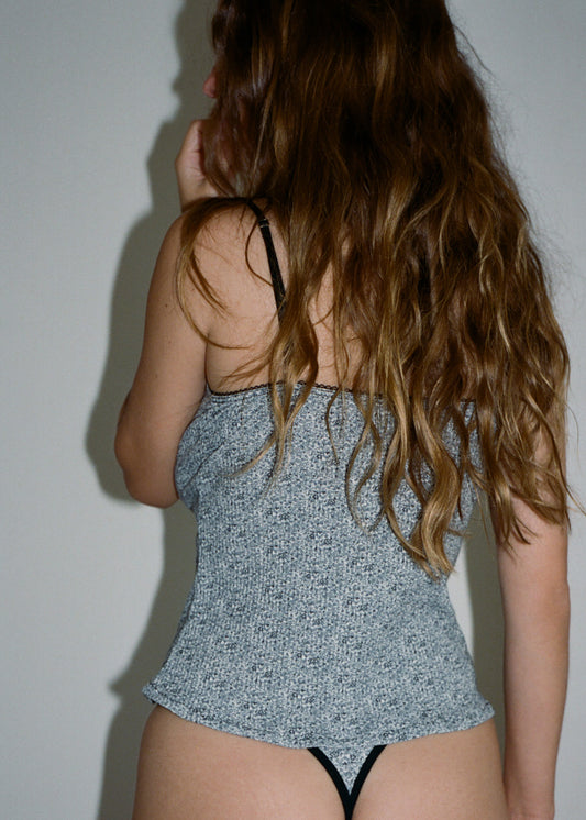 A woman is wearing the Margot Tank in Midnight Floral with the Harri G-string in Midnight Floral by Juem. She has brunette hair, and her left hand is on her chin. She is facing a white wall with her back to the camera. 