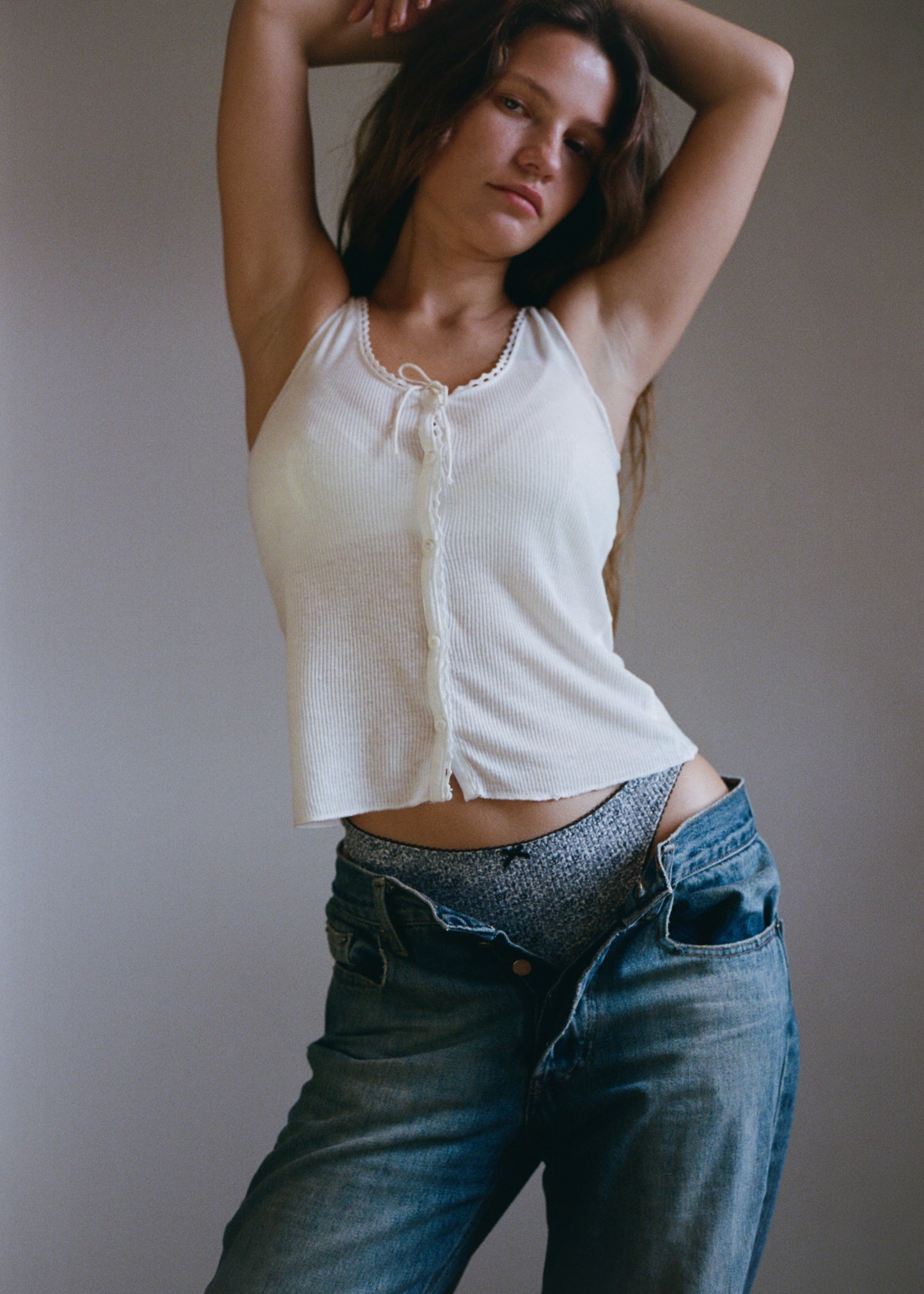 A woman with long brown hair is standing in front of a light coloured wall looking into the camera. She is wearing a white tank top with blue jeans pulled down slightly, showing the Lera Brief in Midnight Floral by Juem. She has her arms raised up, with her hands resting on her head. 