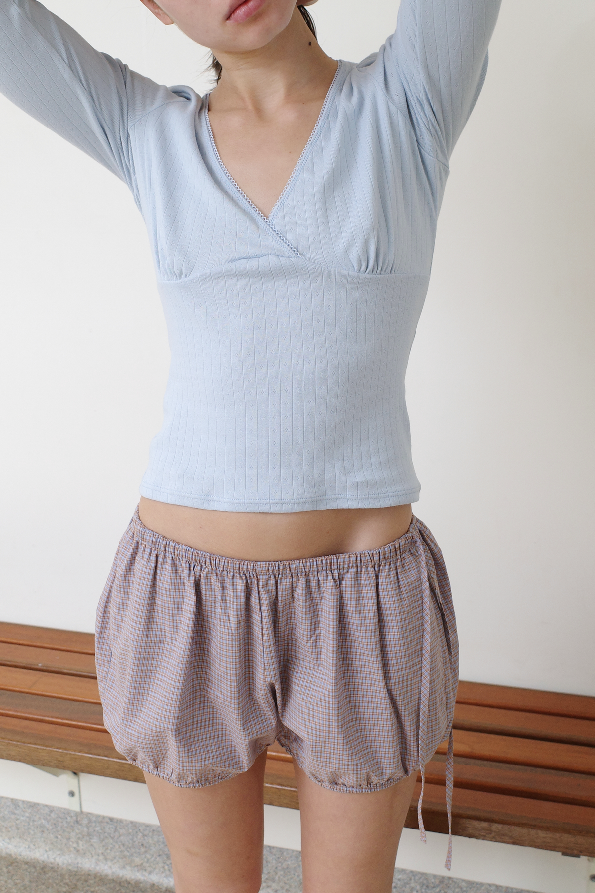A cropped shot of a model with arms raised, wearing the Fern Top - Glacier Blue by Juem, a soft pointelle knit top with a v-neck crossover front and gathered bust panel, styled with the Hazel Bloomer shorts in Blue Tan Check.