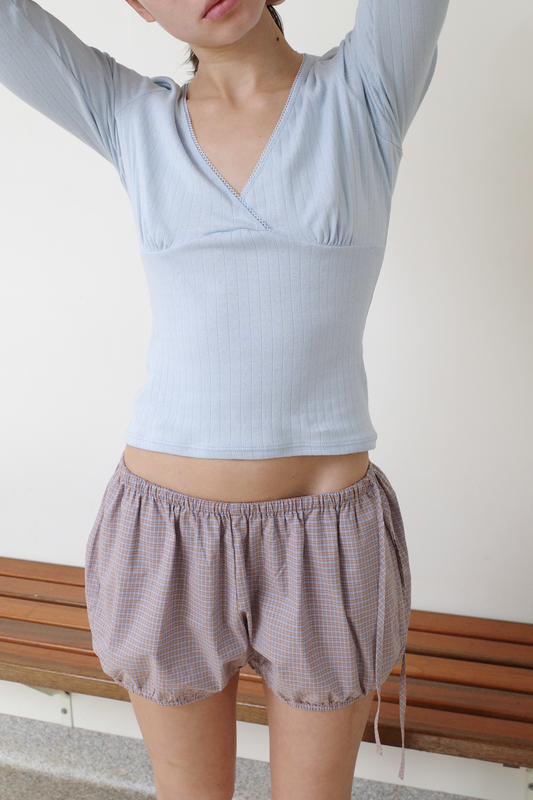A cropped shot of a model with arms raised, wearing the Fern Top - Glacier Blue by Juem, a soft pointelle knit top with a v-neck crossover front and gathered bust panel, styled with the Hazel Bloomer shorts in Blue Tan Check.