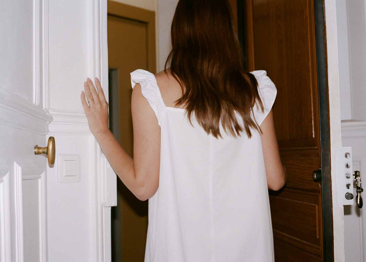 A woman with long brown hair, wearing the Jonni Dress - White by 0, stands in a doorway with her back to the camera—one hand on the doorframe and the other partially visible.