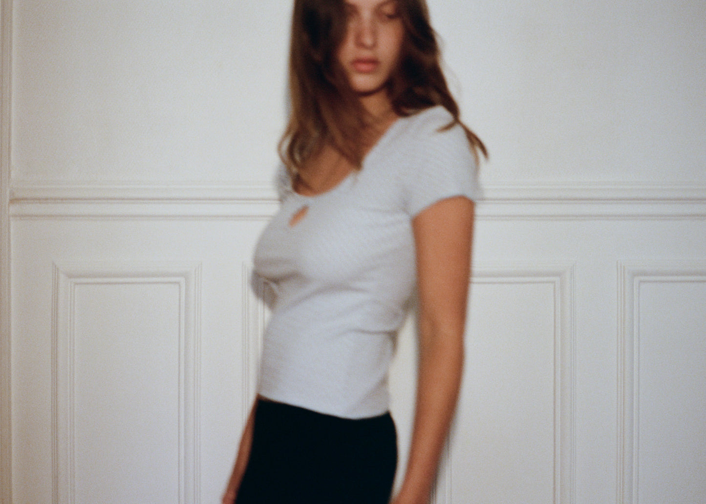 A blurred photo shows a person with long hair in the 0 Effie Tee - Polka, a short-sleeve white ribbed cotton elastane top, and black pants, standing before a white paneled wall. Their face is blurred due to motion.