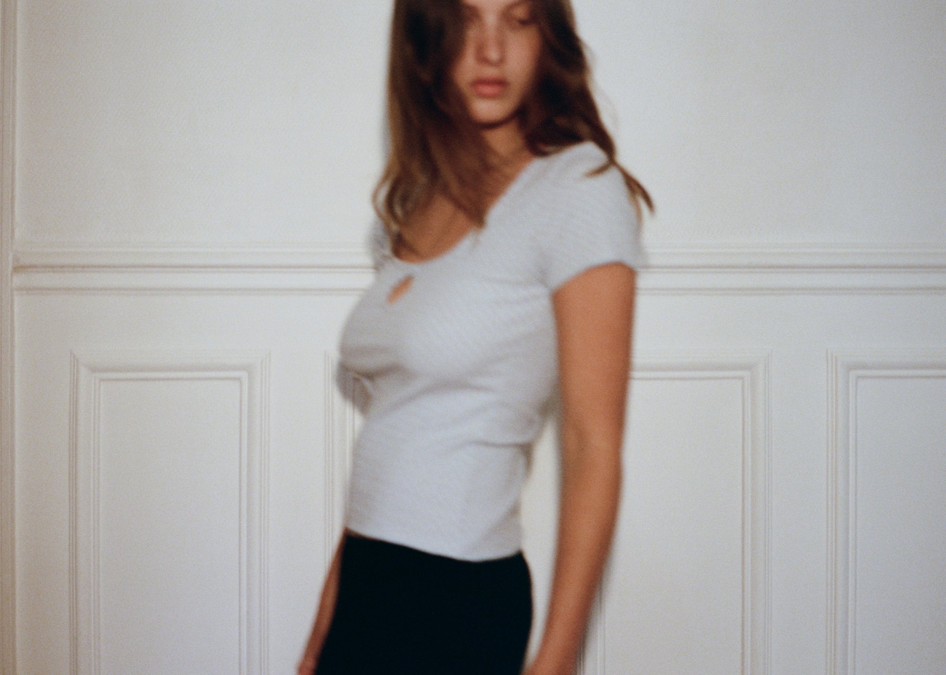 A blurred photo shows a person with long hair in the 0 Effie Tee - Polka, a short-sleeve white ribbed cotton elastane top, and black pants, standing before a white paneled wall. Their face is blurred due to motion.